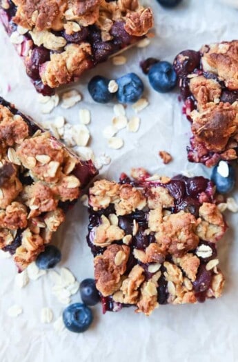 Four breakfast oat bars on a sheet of parchment paper with fresh berries and uncooked rolled oats sprinkled around them