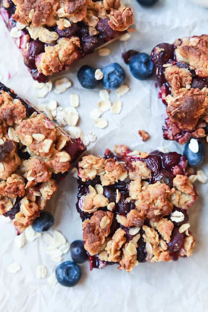 Four breakfast oat bars on a sheet of parchment paper with fresh berries and uncooked rolled oats sprinkled around them