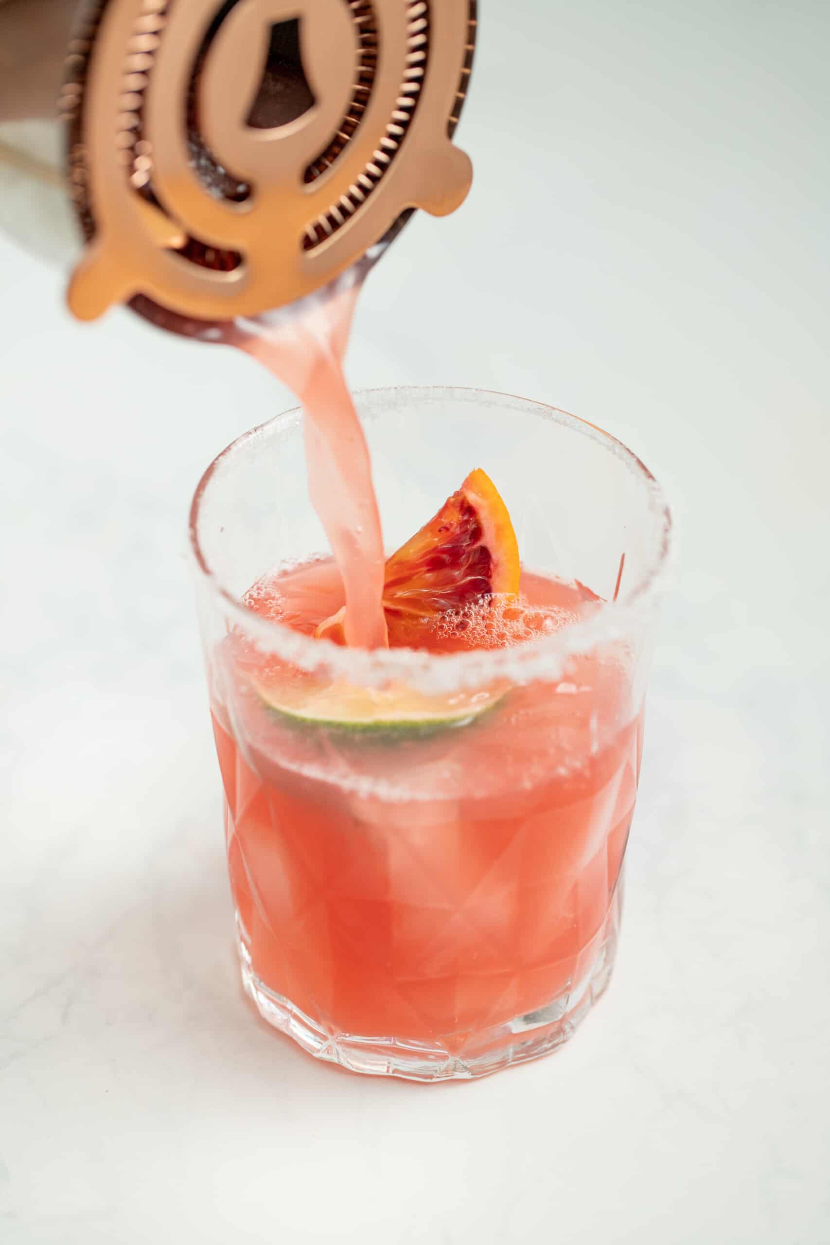 pouring a red colored drinking into a glass with ice, orange wedge, and lime