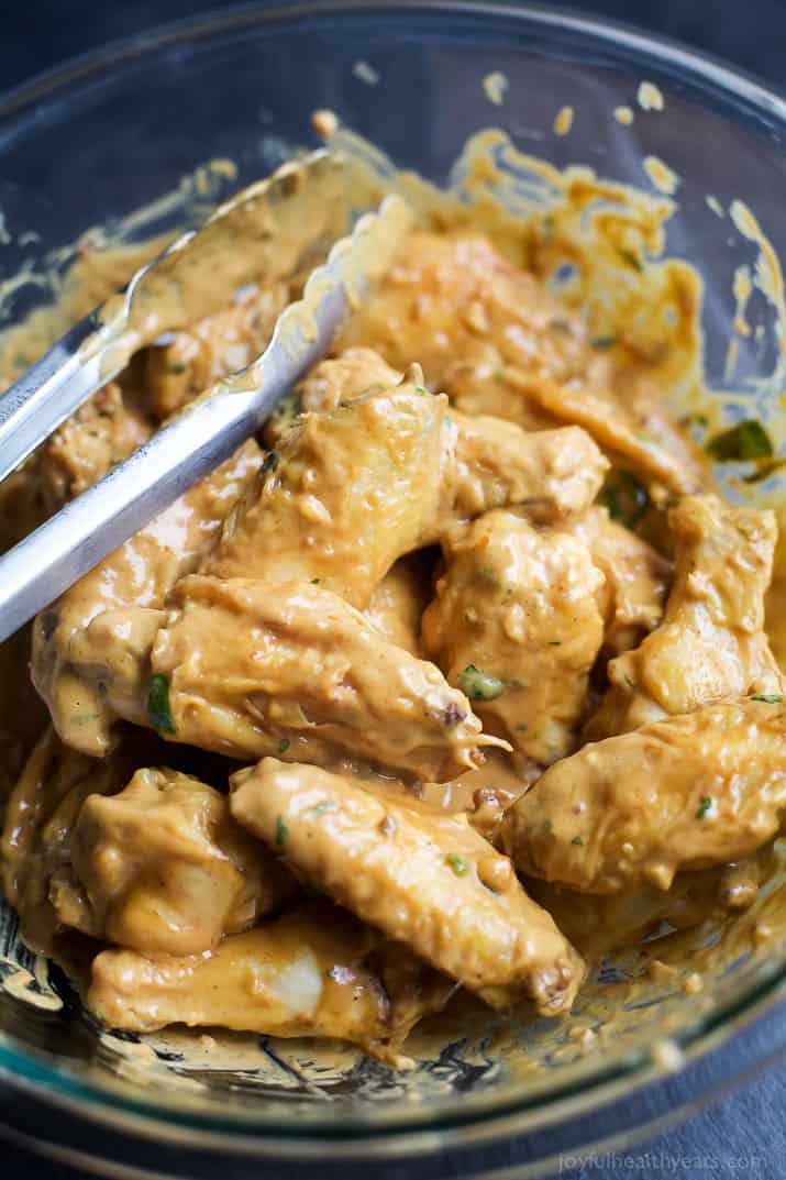 Cooked chicken wings in a glass mixing bowl with peanut sauce and a pair of tongs