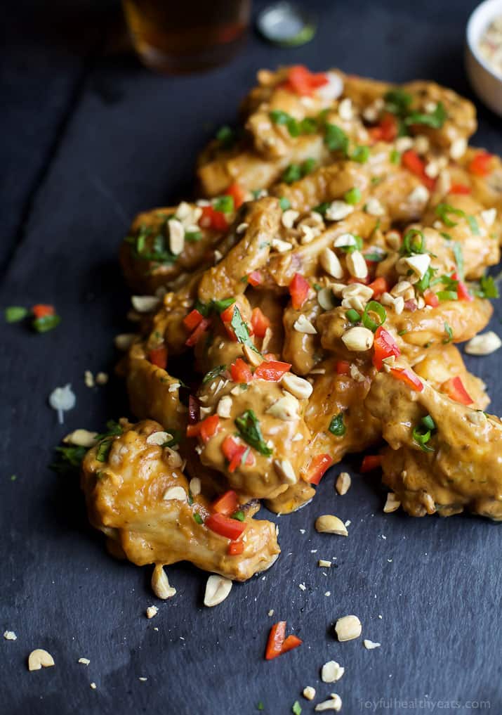 A pile of Thai peanut chicken wings on a table with a bottle of beer and a bowl of peanuts behind them