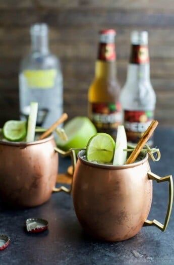 Apple cider mules in copper glasses garnished with cinnamon sticks, apple slices, and lime slices.