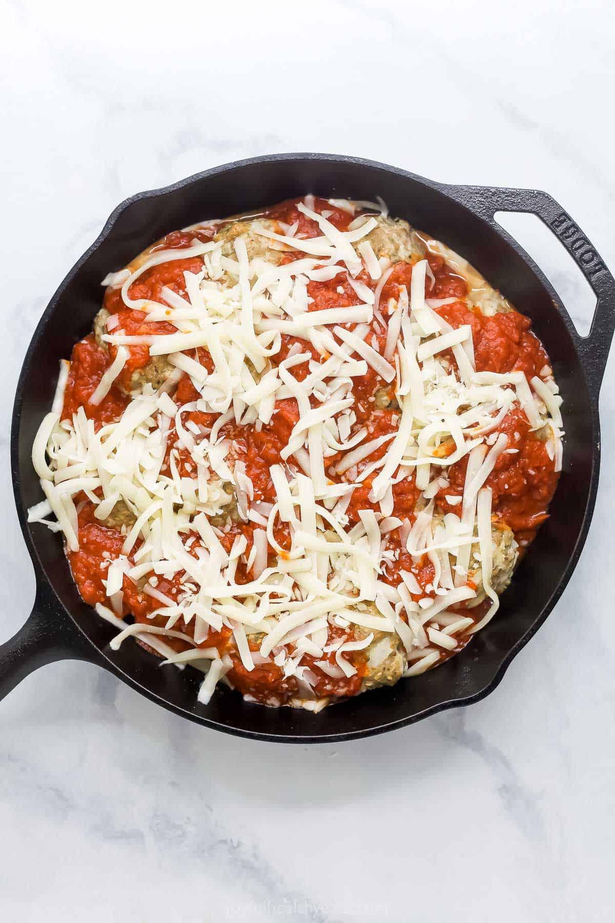 These Baked Turkey Meatballs are cooked in marinara to keep them flavorful and moist, and they’re also loaded with cheese. Done in 30 minutes! Overhead view of marinara and cheese added to skillet of turkey meatballs