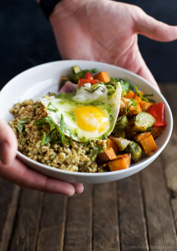 Hands holding a Harvest Roasted Vegetable Grain Bowl topped with an egg