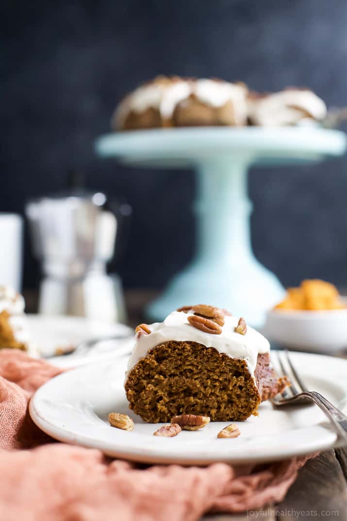 A slice of pumpkin cake on a white plate with the remaining cake on a stand in the background