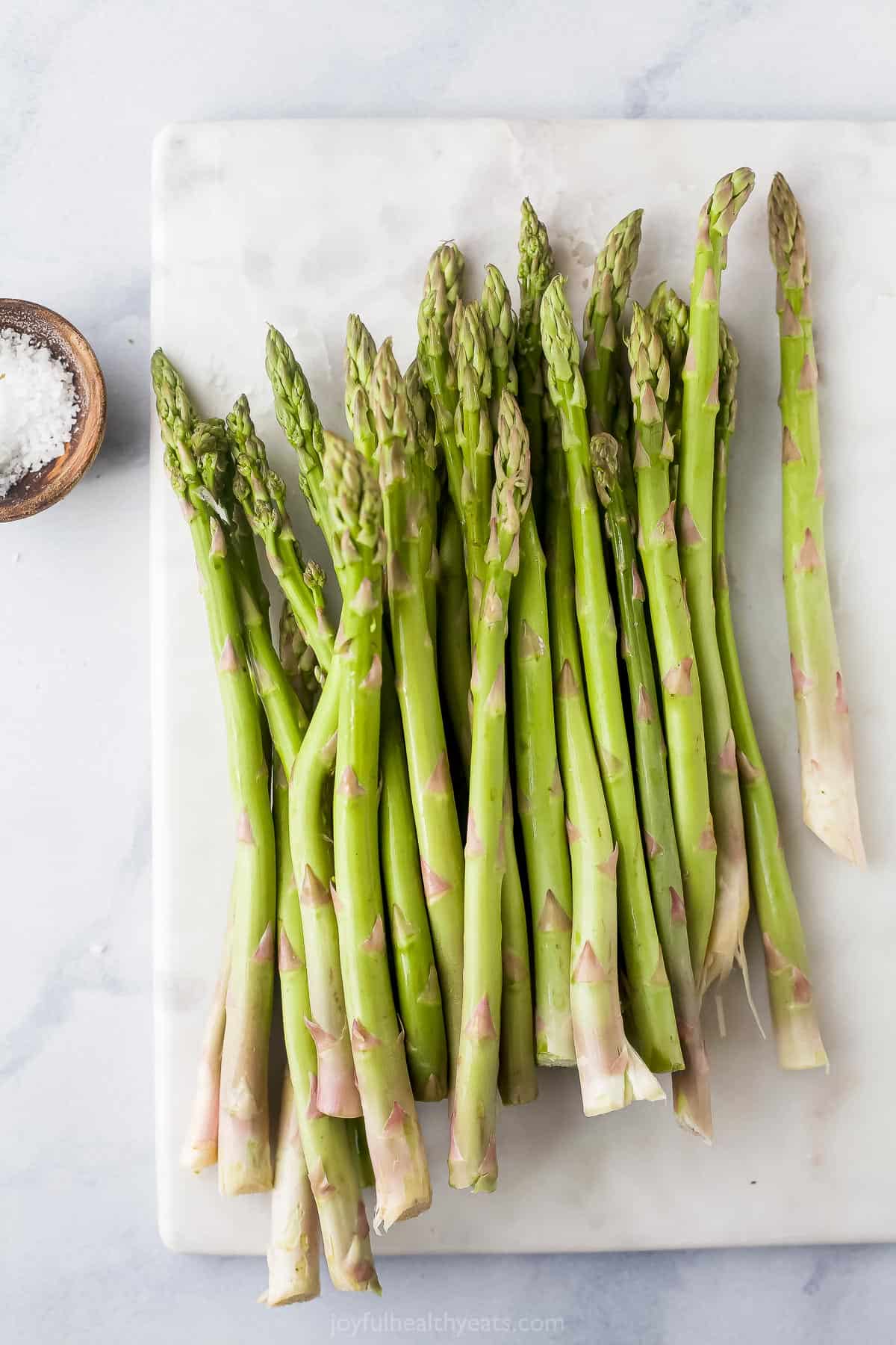Untrimmed stalks of asparagus piled onto a cutting board with a dish of salt beside it