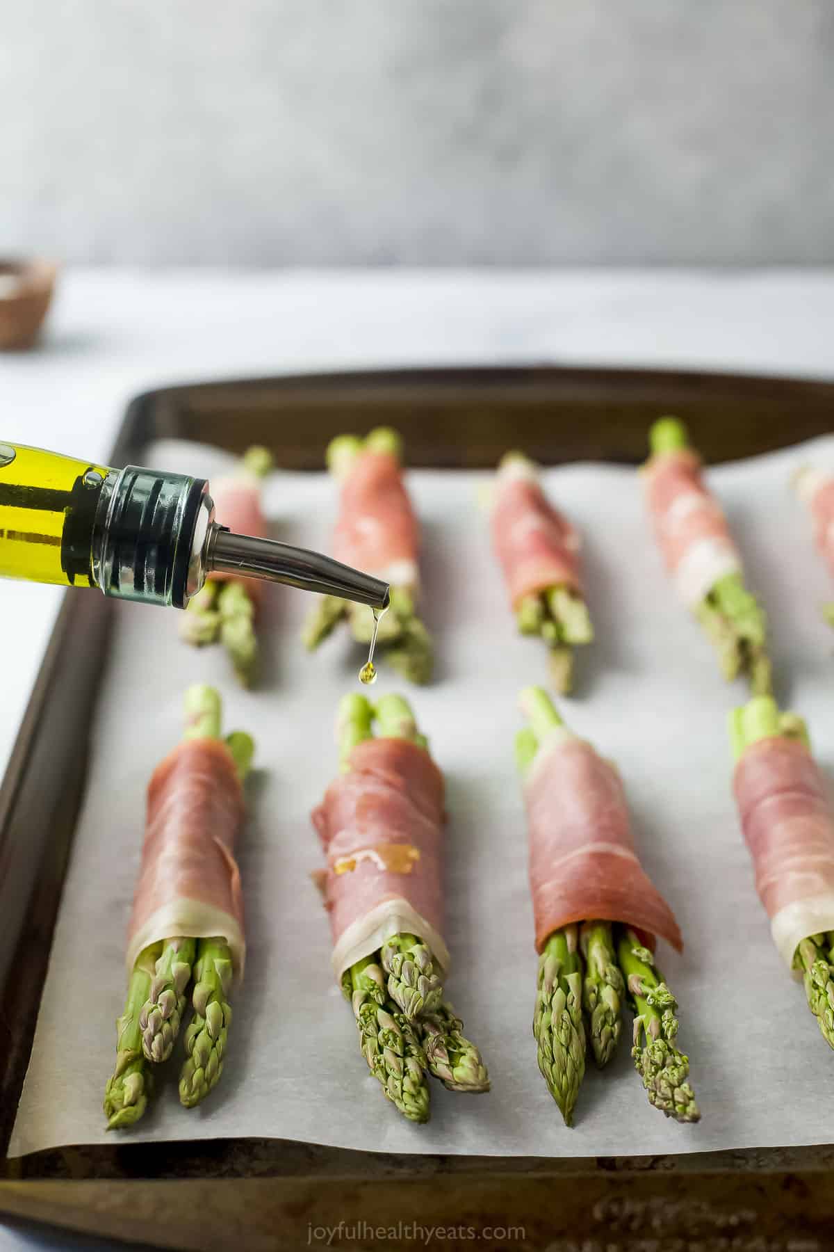 Olive oil being drizzled over prosciutto asparagus bundles on a lined baking sheet