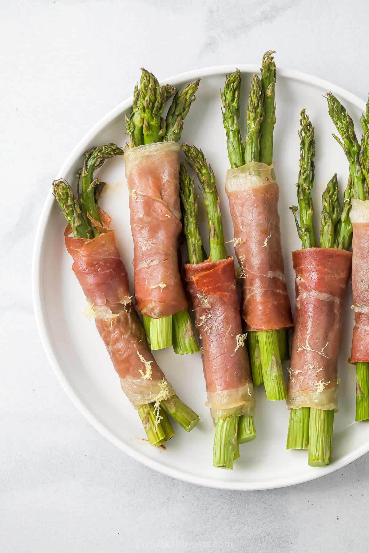 Six prosciutto asparagus wraps on a plate on top of a marble surface