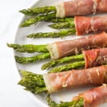 Six prosciutto asparagus wraps lined up on top of a white plate