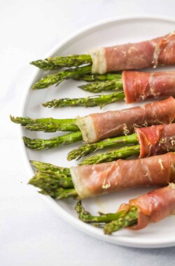 Six prosciutto asparagus wraps lined up on top of a white plate