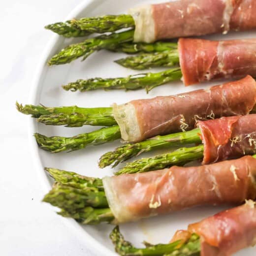 Six prosciutto asparagus wraps lined up on top of a white plate