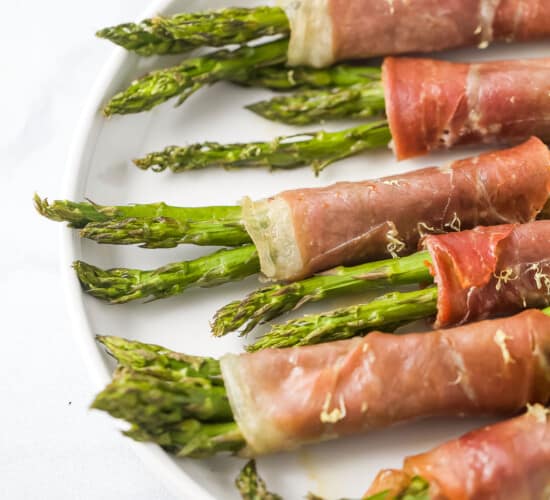 Six prosciutto asparagus wraps lined up on top of a white plate