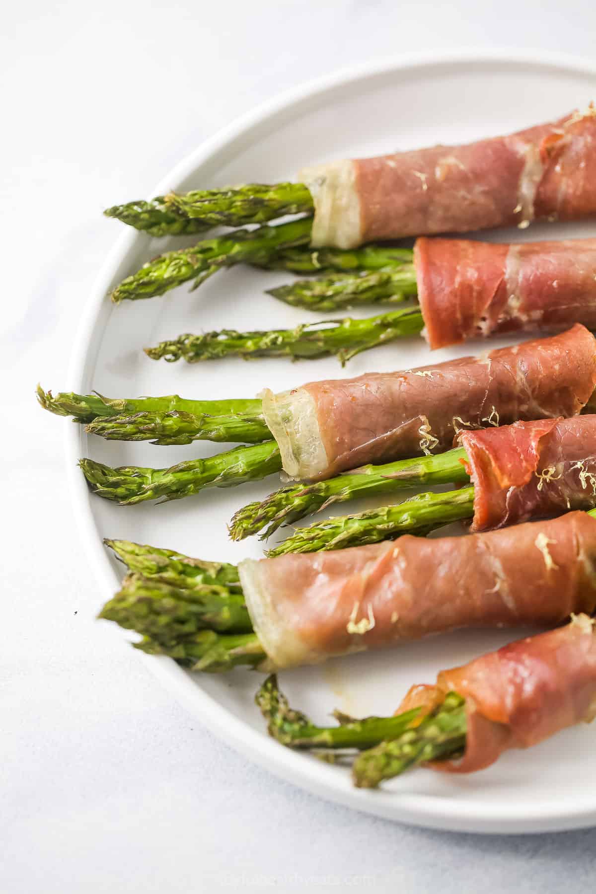 Six prosciutto asparagus wraps lined up on top of a white plate