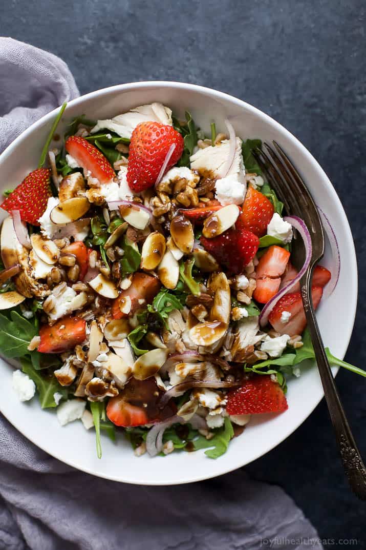 Top view of FARRO ARUGULA STRAWBERRY CHICKEN SALAD in a bowl topped with sliced almonds, goat cheese, and balsamic dressing