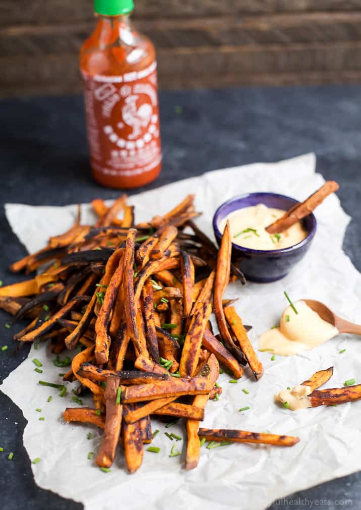 A pile of sweet potato fries on top of a ripped sheet of parchment paper with one fry being dipped into a bowl of aioli.