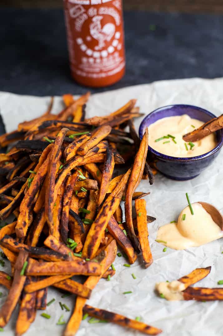 A pile of freshly-baked fries beside a dish of aioli dipping sauce with a sriracha bottle behind them