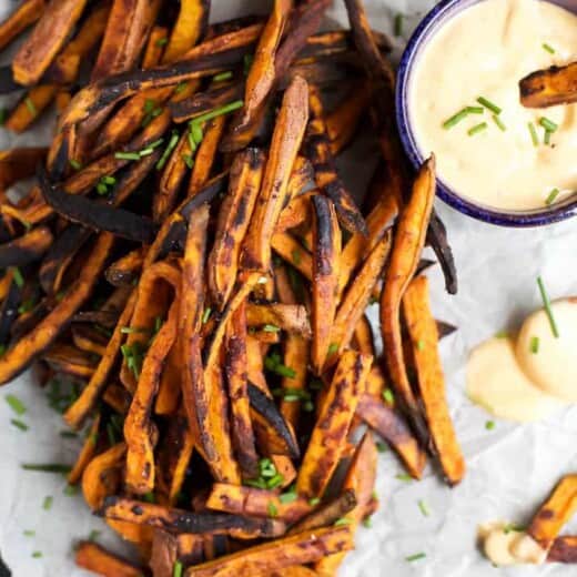 An overhead view of a batch of sweet potato fries on a sheet of parchment paper with honey sriracha sauce beside them