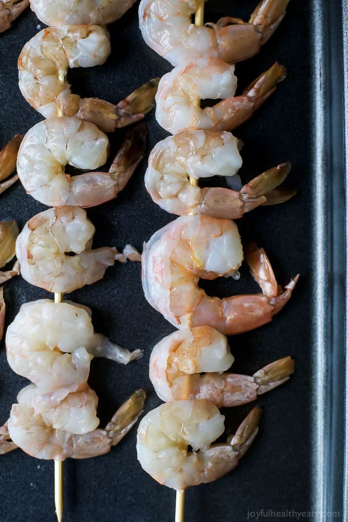 A bunch of raw shrimp threaded through two wooden skewers on top of a cast-iron cookie sheet