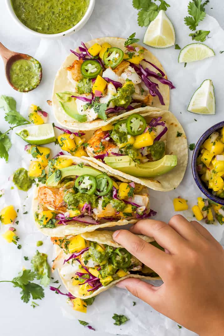 Overhead view of a hand grabbing a fish taco with mango salsa, avocado and garnishes