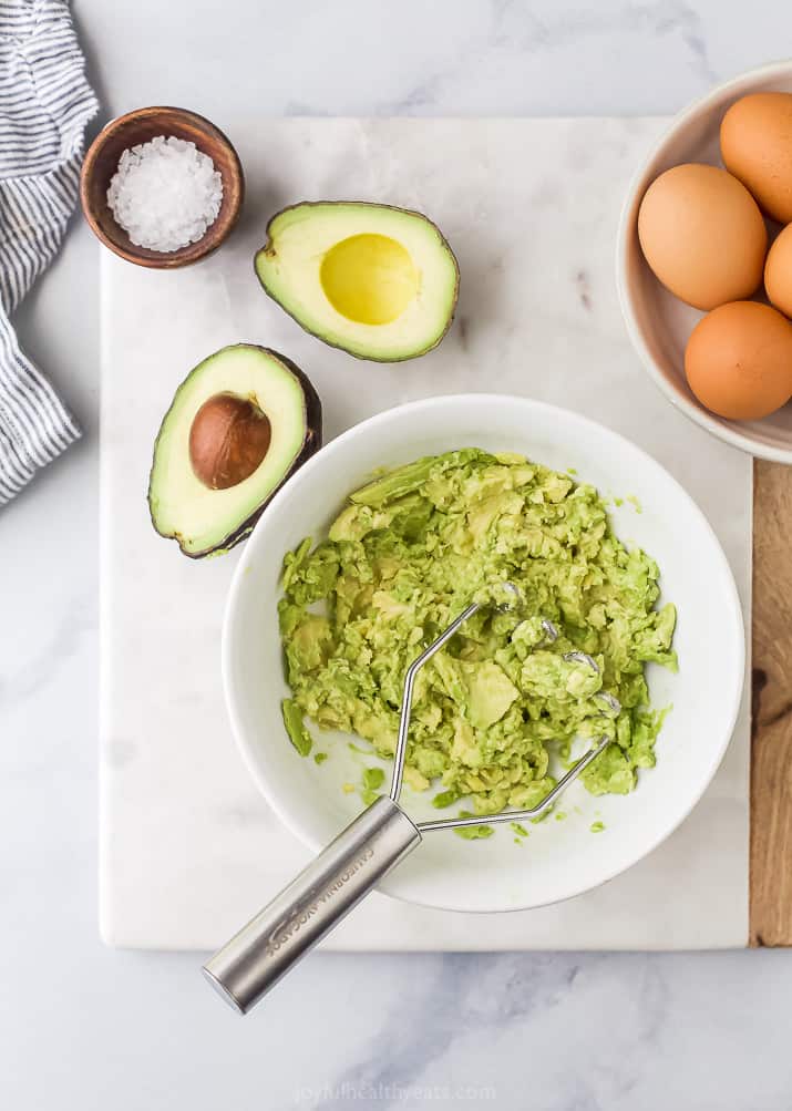 Overhead view of smashed avocado in a bowl next to a halved avocado, a bowl of salt, and a bowl of eggs