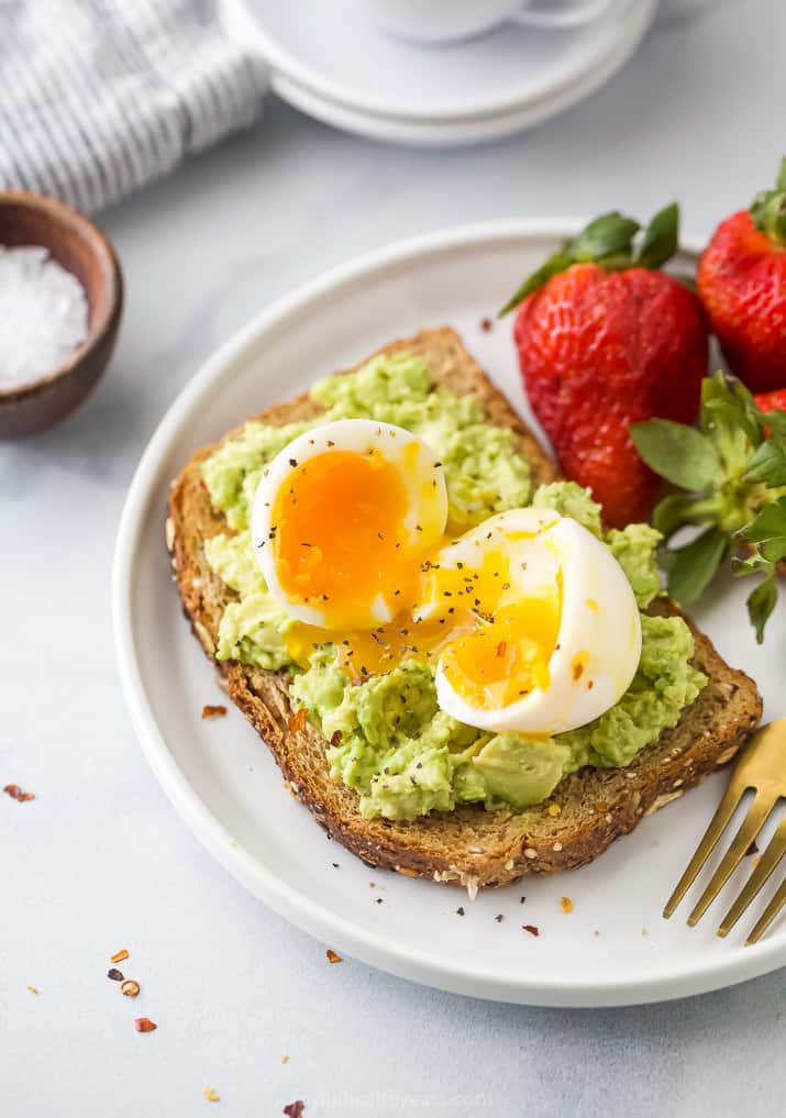 Overhead view of a plate of smashed avocado toast topped with a soft boiled egg served with fresh strawberries