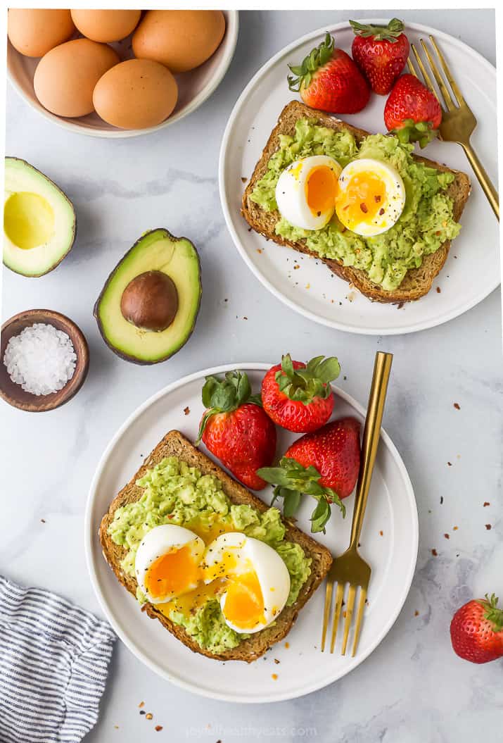 Overhead view of two plates of smashed avocado toast topped with a soft-boiled egg and served with fresh strawberries