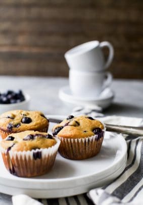 A plate holding three homemade muffins with a stack of two coffee cups in the background