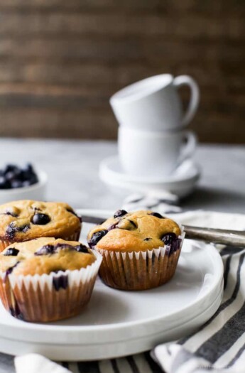 A plate holding three homemade muffins with a stack of two coffee cups in the background