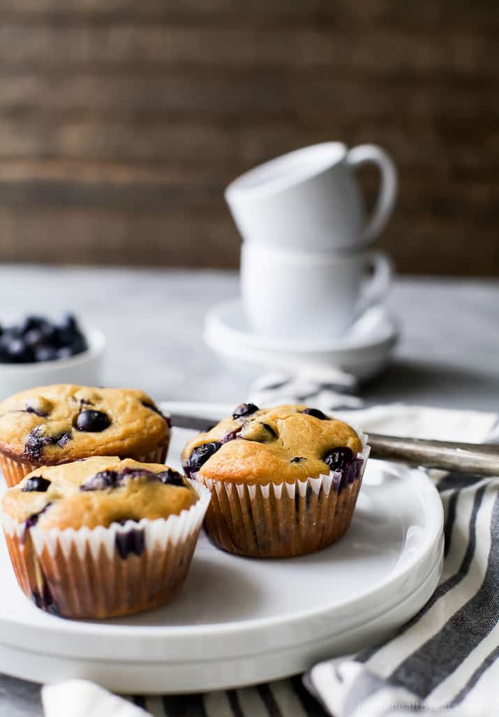 A plate holding three homemade muffins with a stack of two coffee cups in the background