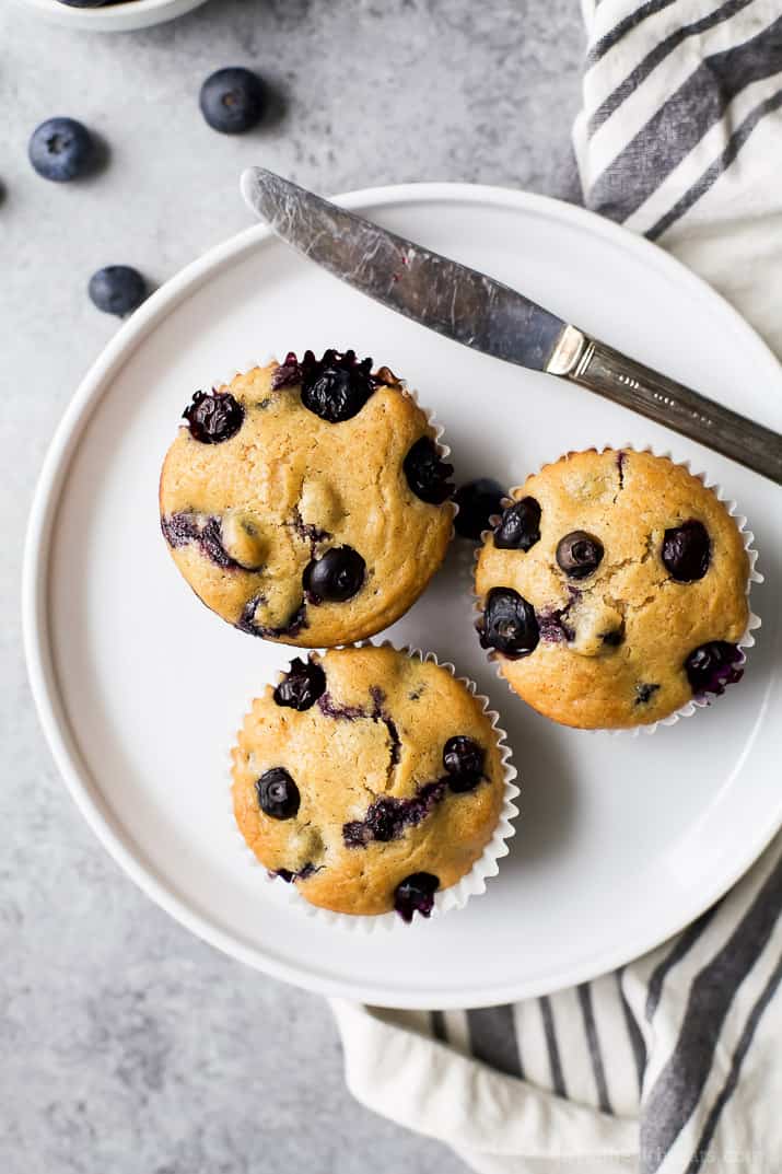 Three moist blueberry muffins on a plate with a butter knife beside them