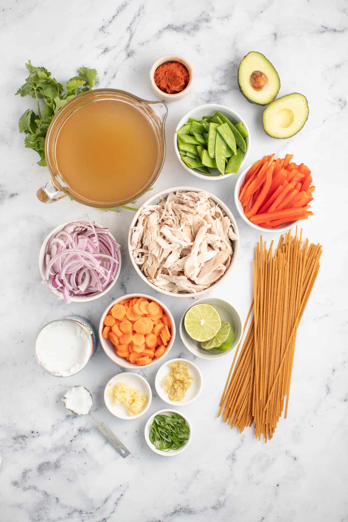 an assortment of ingredients on a countertop in small prep bowls like broth, sliced onions, snap peas, sliced peppers, carrots, limes, garlic, ginger, and noodles