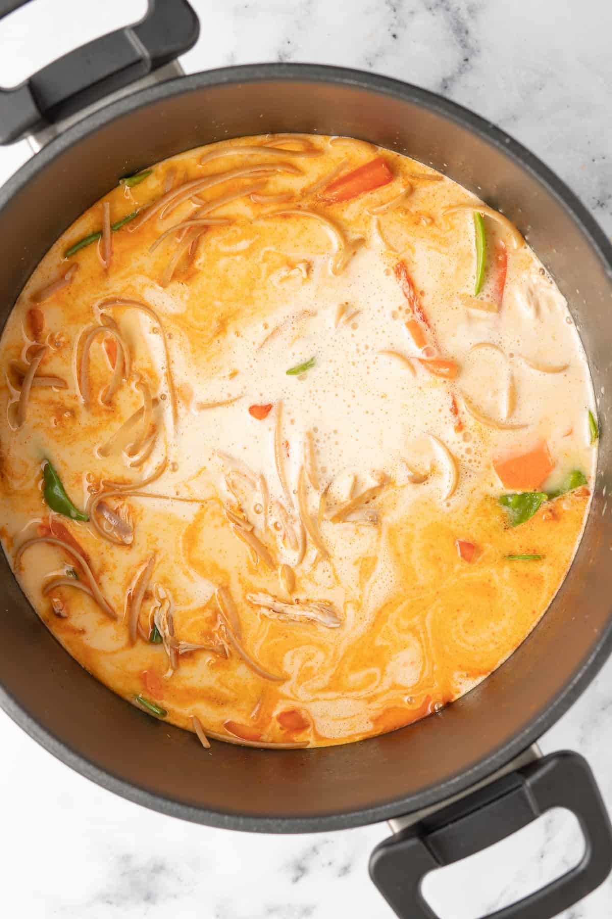 a pot of chicken thai soup with an orange broth, fresh herbs, vegetables, and noodles