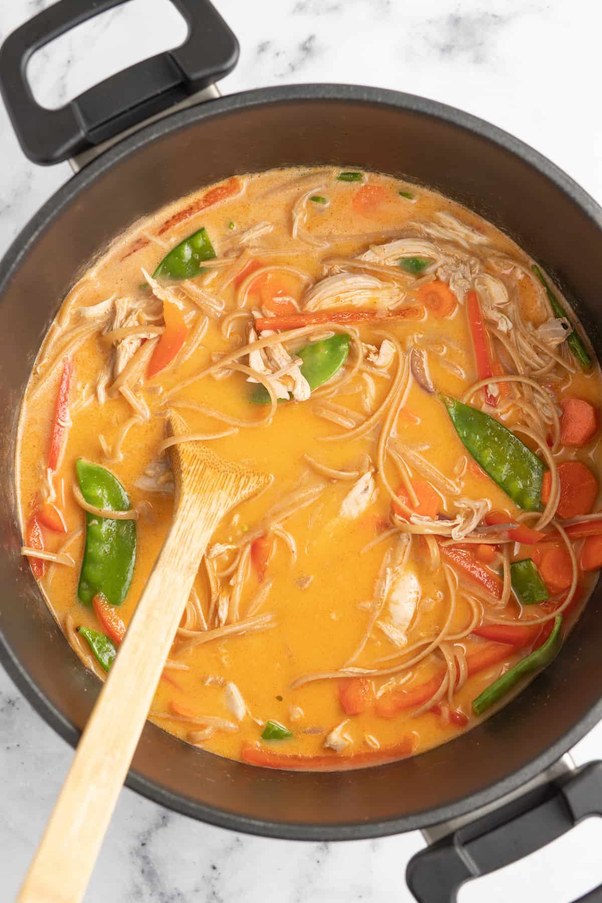 a pot of chicken thai soup with an orange broth, fresh herbs, vegetables, and noodles