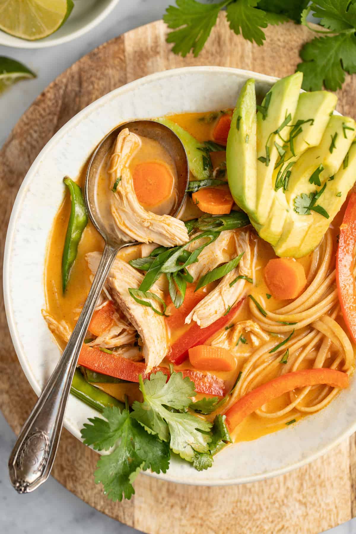 thai chicken noodle soup with an orange colored broth, rice noodles, avocados, and fresh herbs with a large metal spoon
