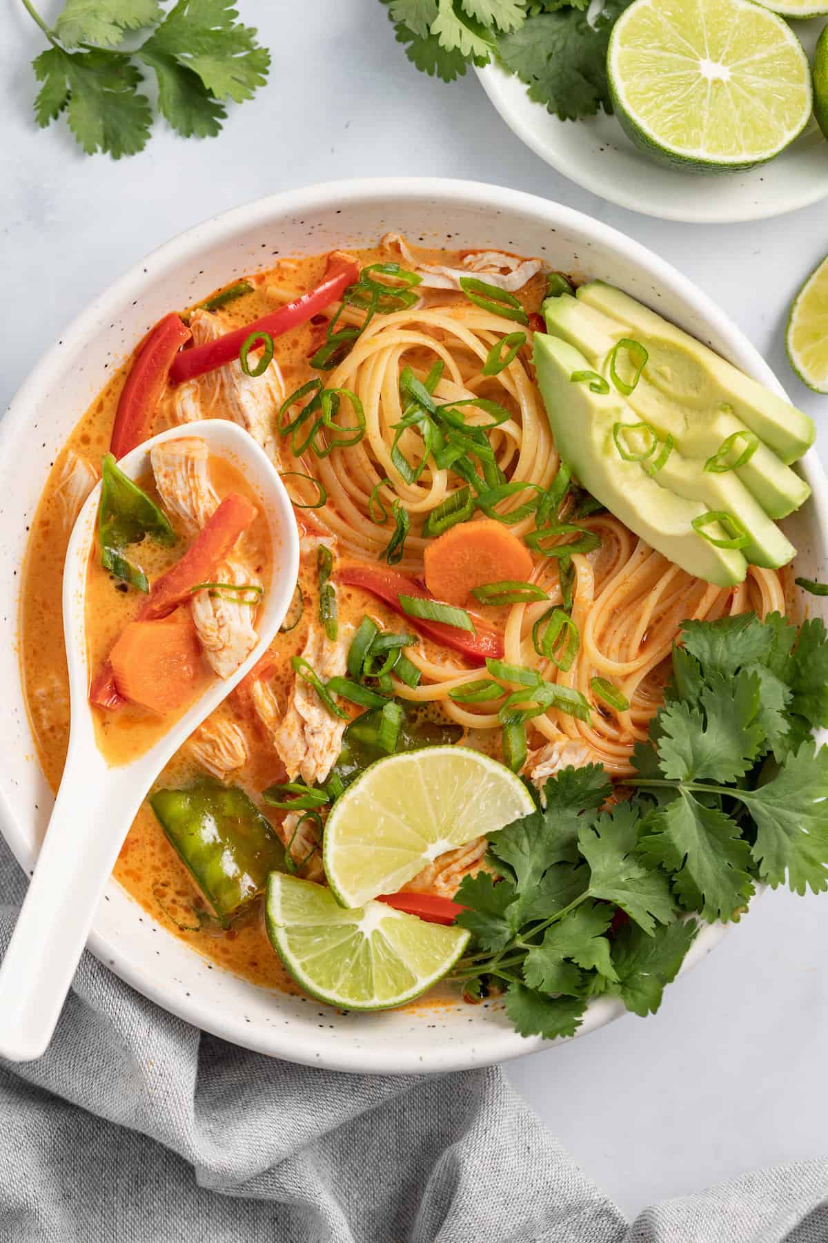 thai chicken noodle soup with an orange colored broth, rice noodles, avocados, and fresh herbs with a ladle s،