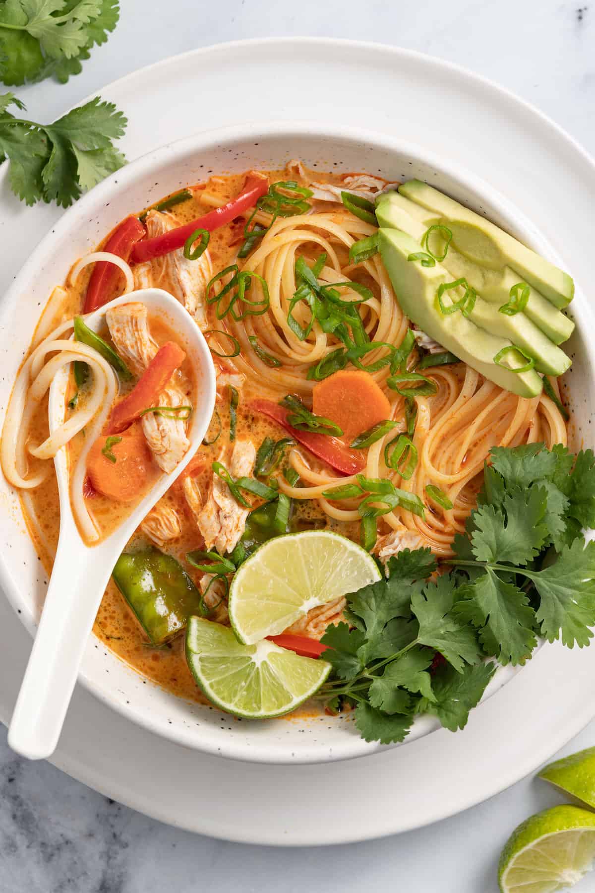 thai chicken noodle soup with an orange colored broth, rice noodles, avocados, and fresh herbs