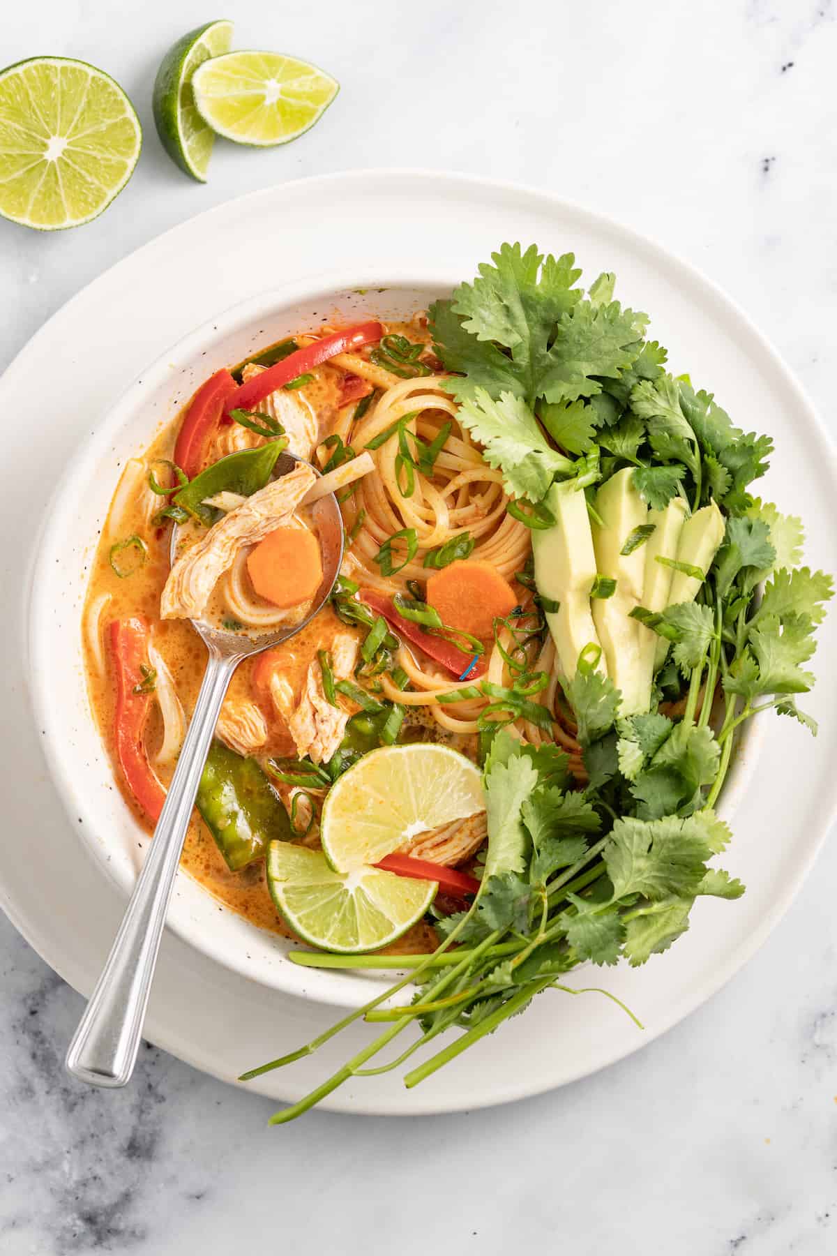 thai chicken noodle soup with an orange colored broth, rice noodles, avocados, and fresh herbs