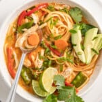thai chicken noodle soup with an orange colored broth, rice noodles, avocados, and fresh herbs