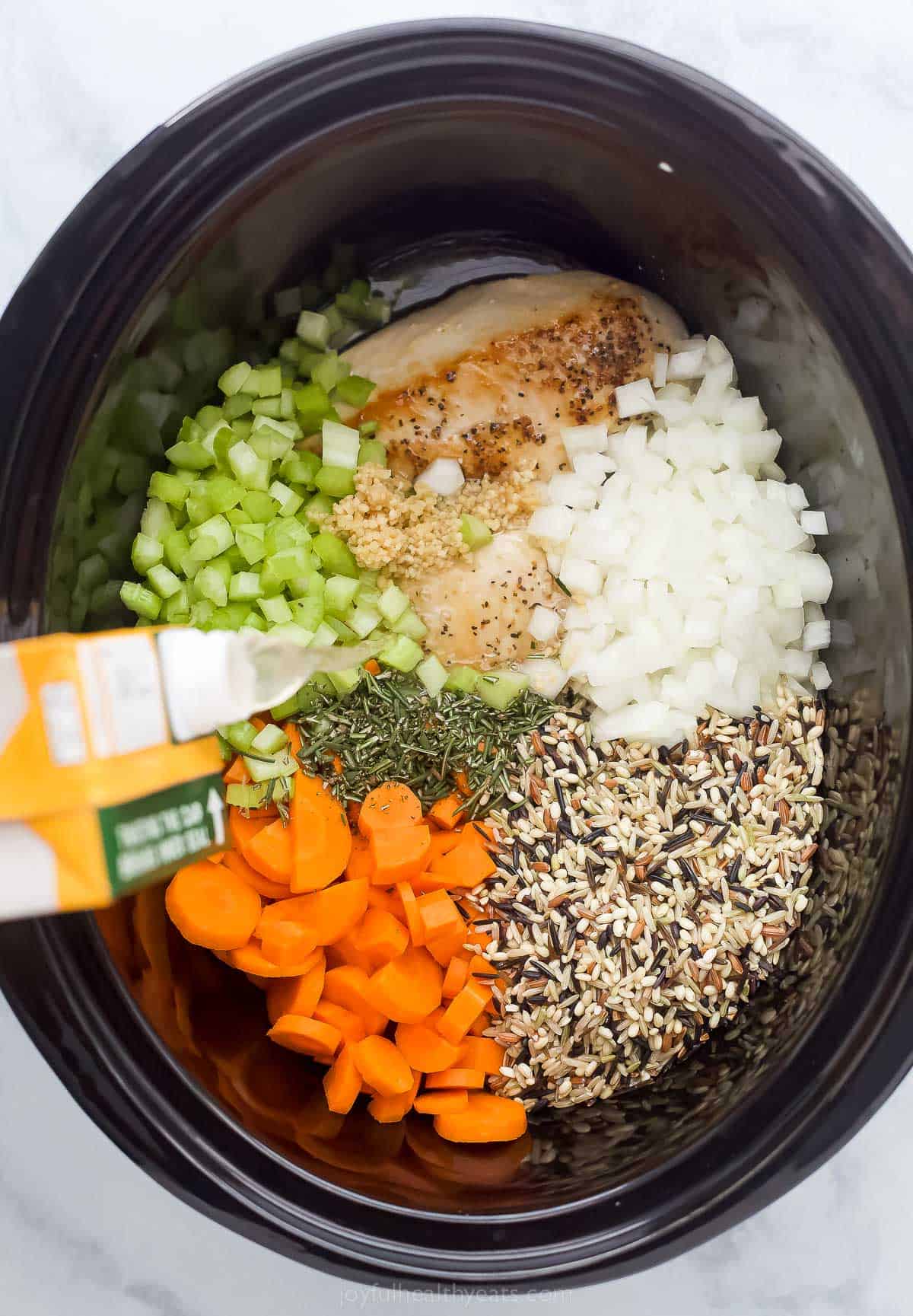 pouring chicken broth into a crockpot with vegetables, chicken, and wild rice