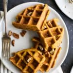A waffle broken into four sections on a plate with a few chopped pecans and a fork