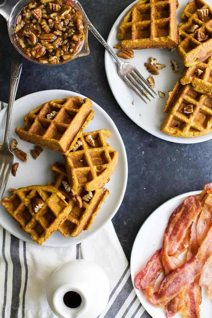 A table shown from above with waffles, bacon, syrup, cloth napkins and a bowl of pecans on it