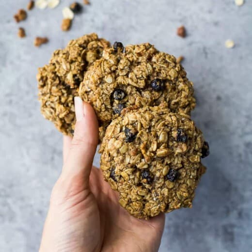 A hand holding three blueberry oatmeal cookies above a kitchen countertop