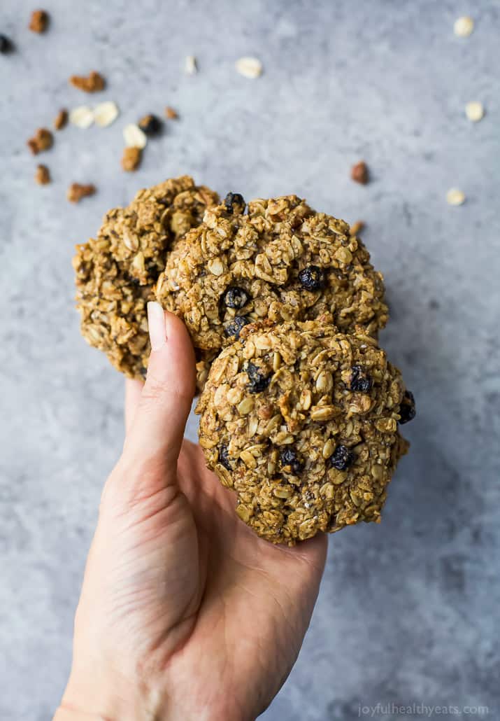 A hand holding three blueberry oatmeal cookies above a kitchen countertop