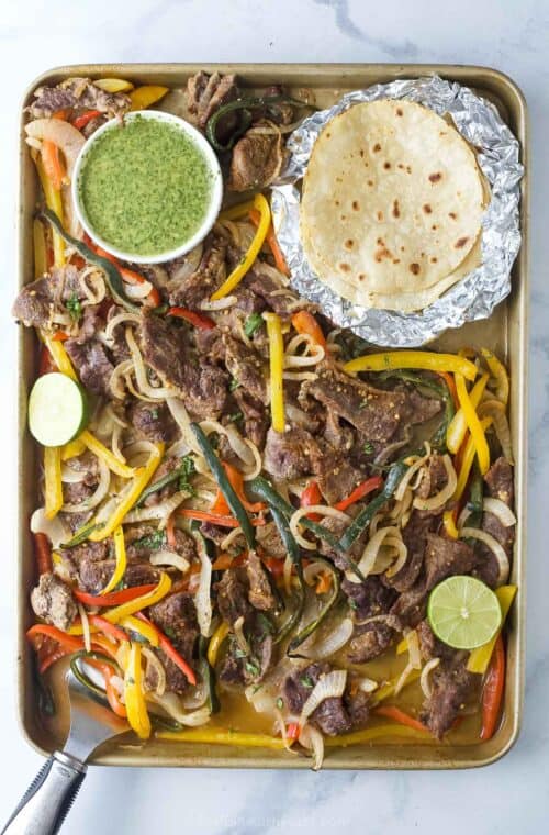 Sheet pan steak fajitas with cilantro chimichurri and flour tortillas on the side.