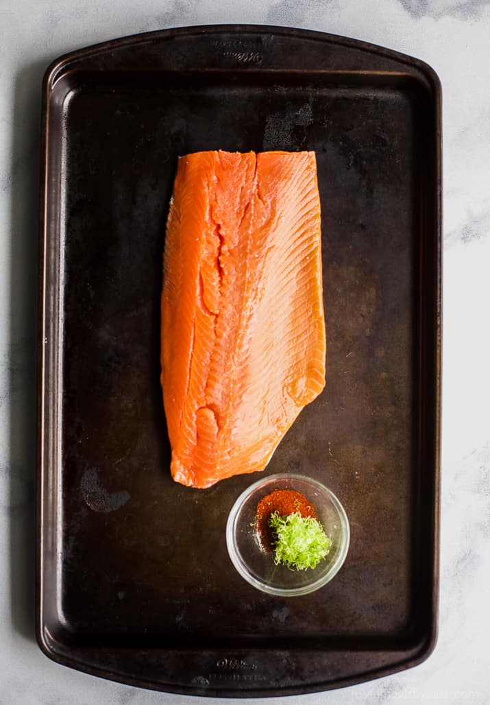 Raw salmon fillet on a baking sheet next to a cup of spices