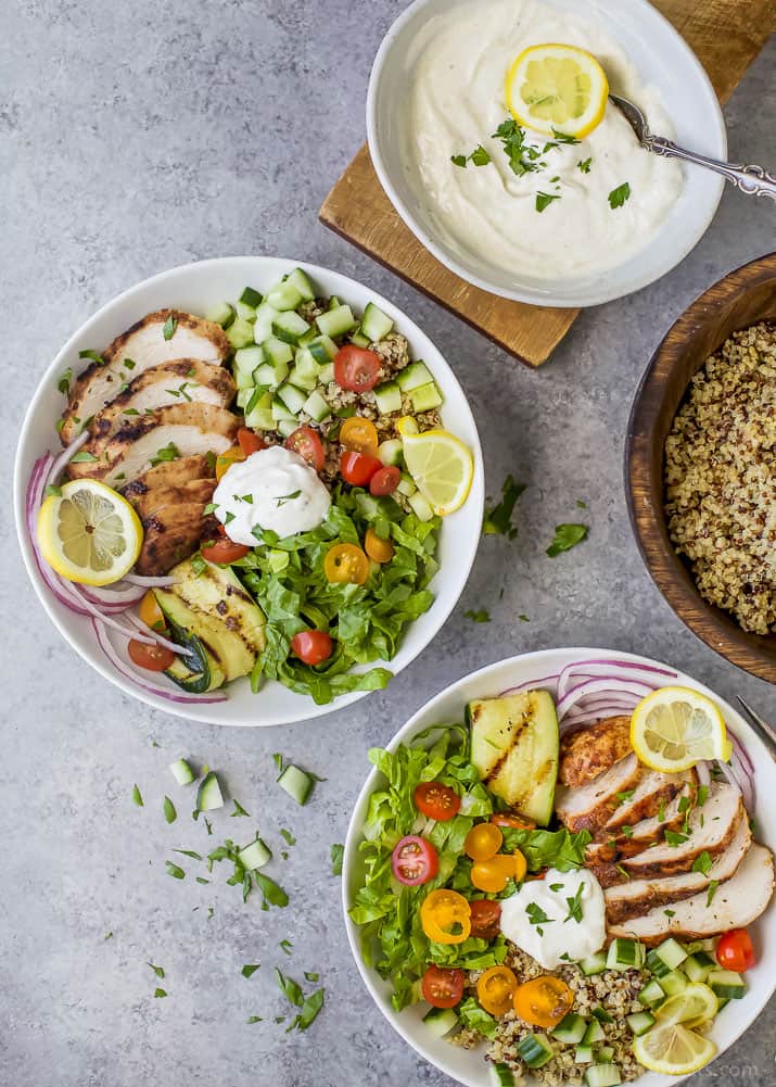 Two chicken shawarma bowls on a countertop beside a bowl of yogurt sauce and a bowl of quinoa