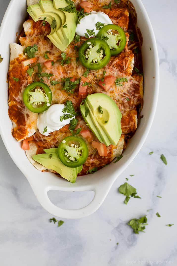 This saucy sweet potato black bean enchiladas recipe with smokey seasonings, spicy salsa, and melty cheese is an easy Tex-Mex dinner. Angled photo of baked enchiladas with cilantro, chopped chili, and avocado.