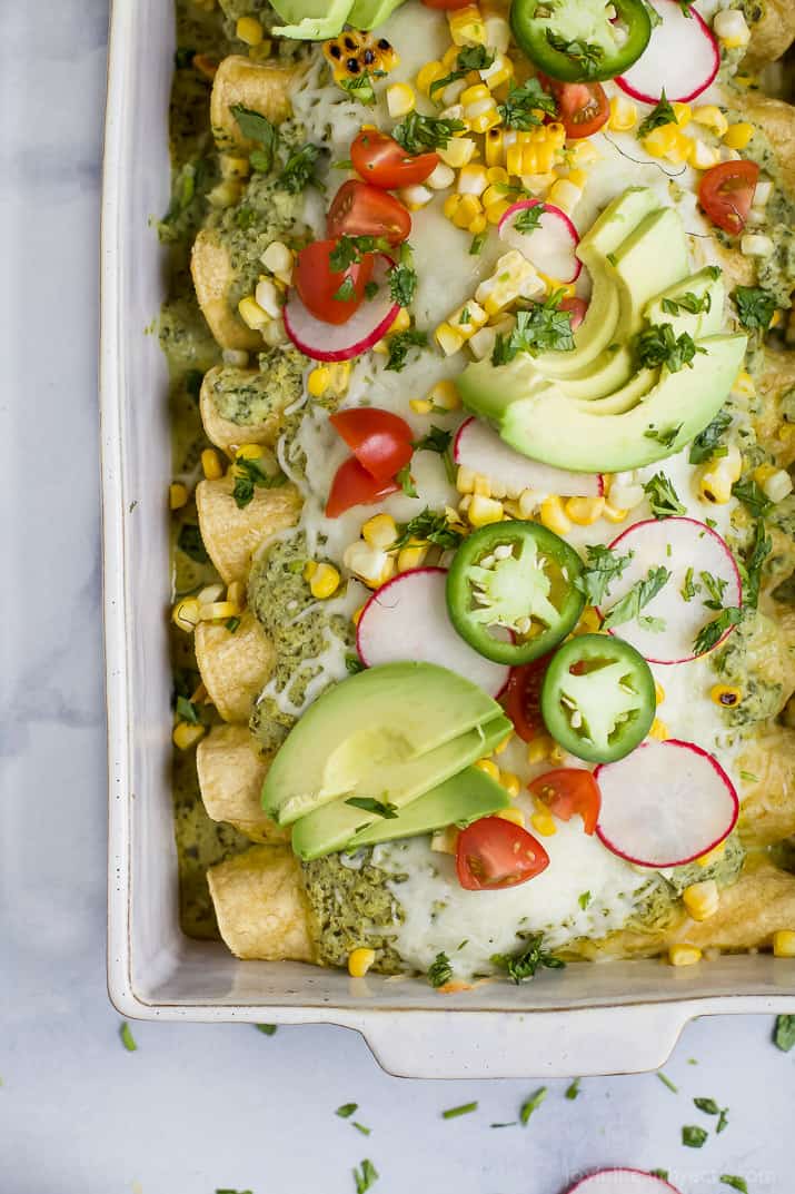 Top view of Chicken Enchiladas in a baking dish topped with Poblano Cream Sauce, fresh avocado, tomato, corn and radishes