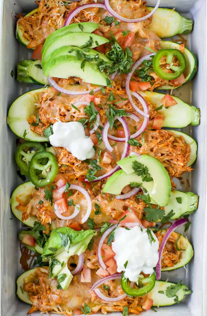 Top view of Chicken Enchilada Zucchini Boats in a baking dish topped with avocado, red onion, sour cream and fresh herbs