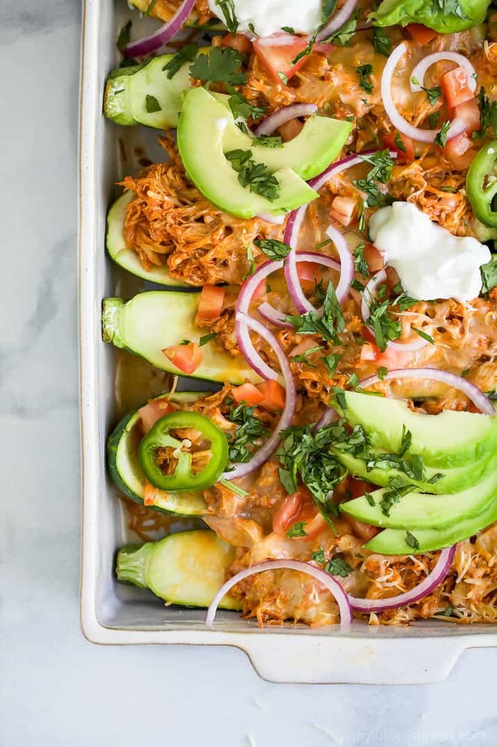 Top view of Chicken Enchilada Zucchini Boats in a baking dish topped with avocado, red onion, sour cream, and fresh herbs